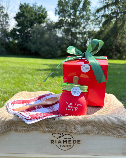 A Sweet Apple Harvest Simmer Kit by Riamede Farm is displayed on a wooden crate outdoors. The kit includes a red jar with a green lid and wooden spoon, a red gift box tied with a green ribbon, and a red-and-white checkered cloth napkin.